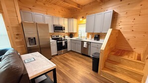 A compact kitchen with wooden cabinets, a refrigerator, microwave, stove, and a sink. There's a small table and a trash bin. - Brand new cabin with HOT TUB, Dock & Lake Access (Butler)