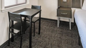 Una habitación de hotel con cama, escritorio con silla, mesita de noche, luz de pared y ventana con persianas.