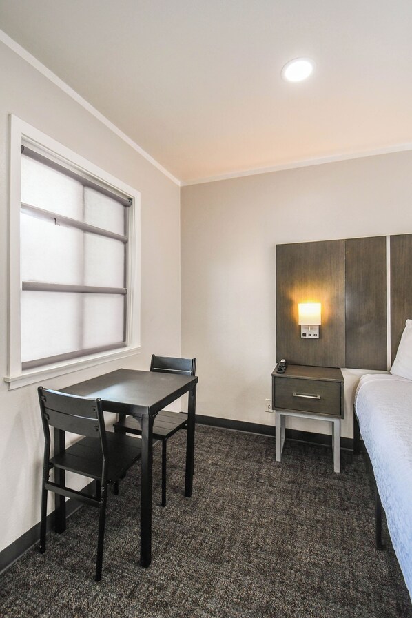 A hotel room with a bed, a desk with a chair, a nightstand, a wall-mounted light, and a window with blinds.