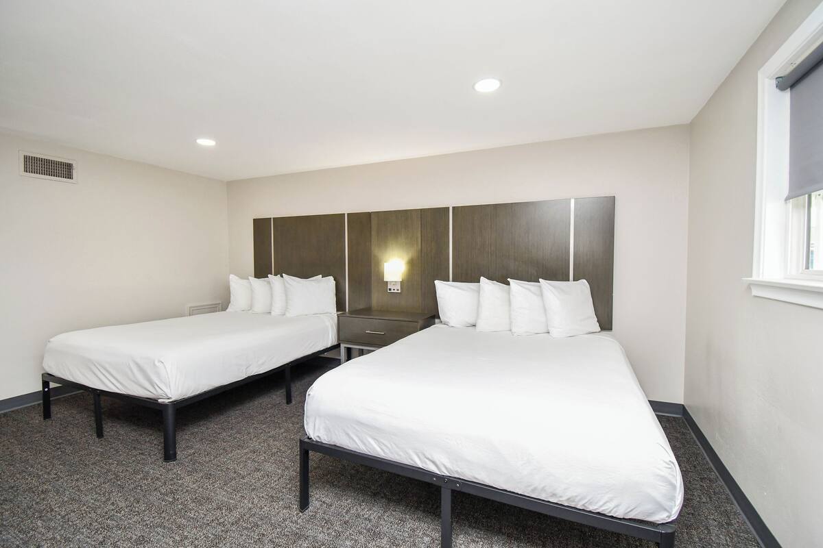 A hotel room with two single beds, a nightstand, a wall-mounted light, and a window with a blind.