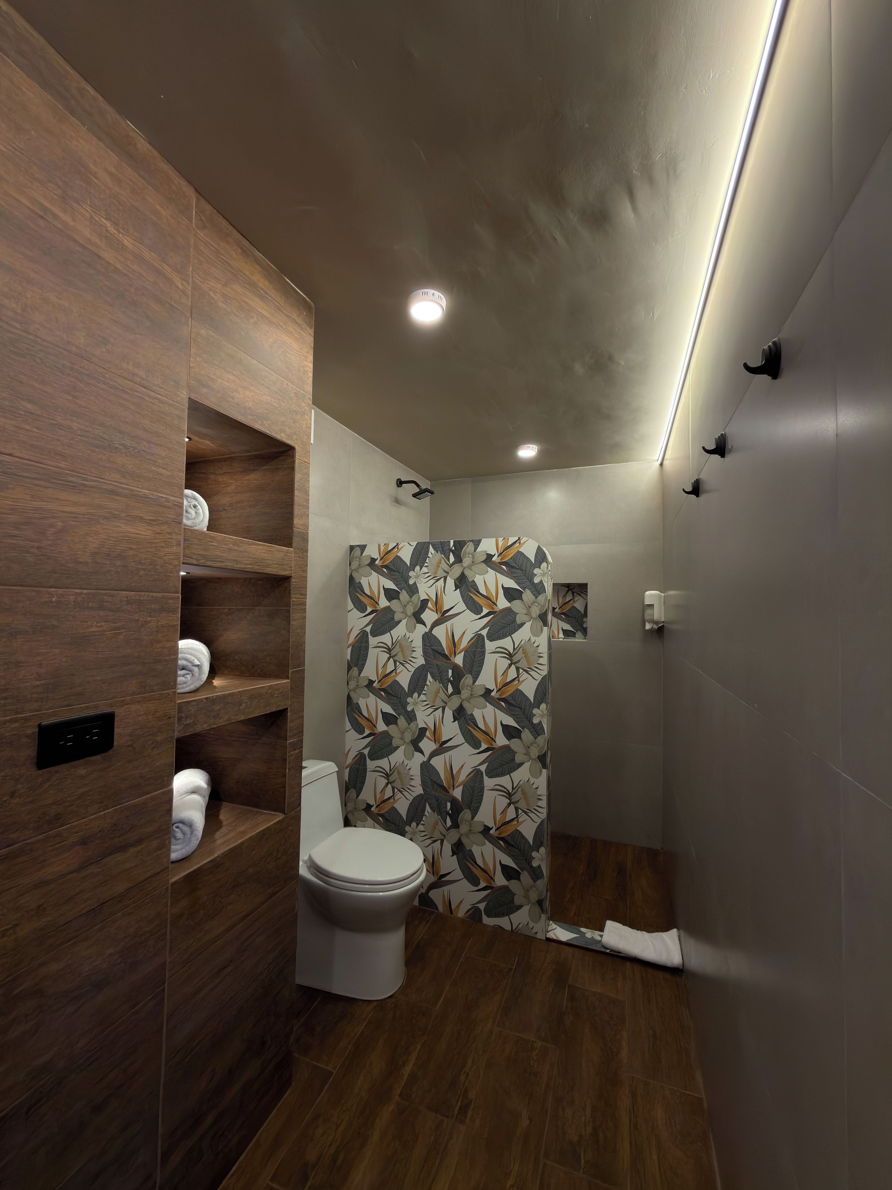 A modern bathroom with wooden shelves, a toilet, a shower with a floral shower curtain, and recessed ceiling lights.