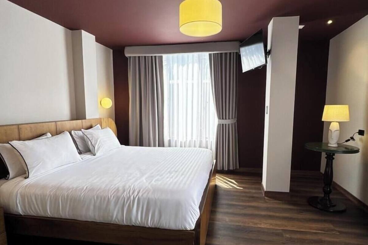 Habitación de hotel con una cama grande, suelo de madera, televisor montado en la pared y una lámpara sobre una mesita.
