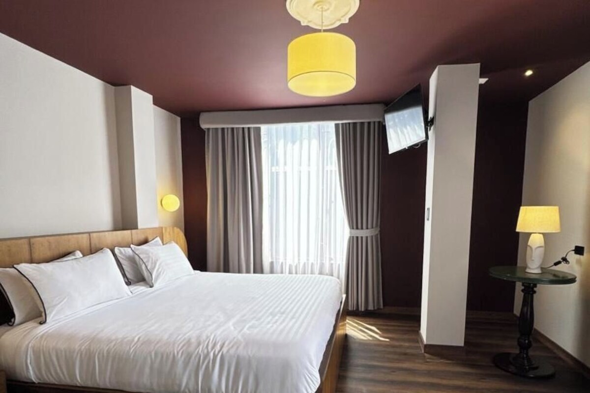 Habitación de hotel con una cama grande, suelo de madera, televisor montado en la pared y una lámpara sobre una mesita.