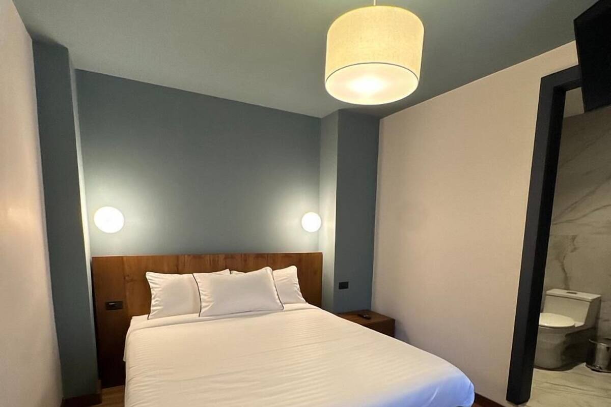 Una habitación de hotel moderna con una cama grande, cabecera de madera, dos lámparas de noche y una lámpara de techo redonda.