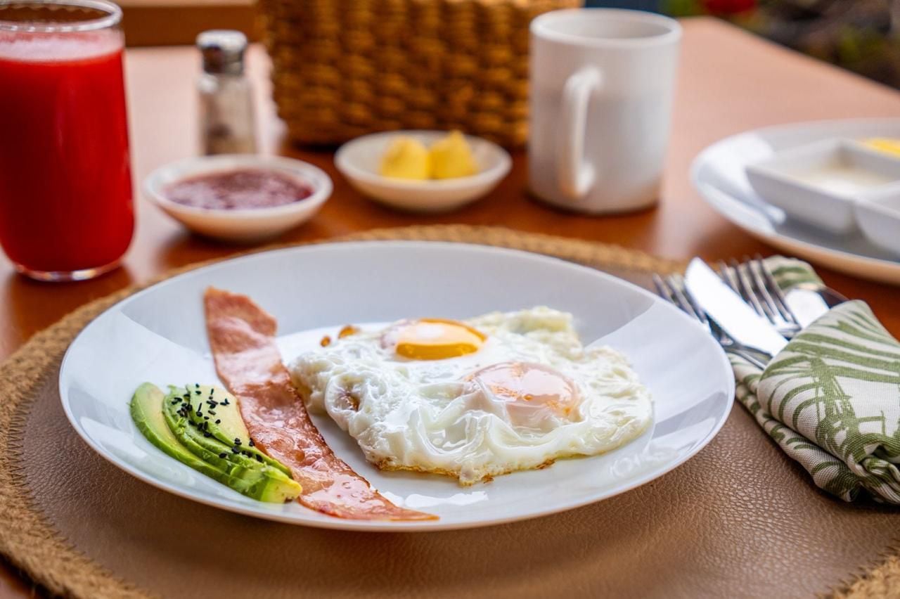 Un plato de desayuno con huevos, aguacate y tocino, un vaso de jugo rojo y una taza blanca sobre una mesa.
