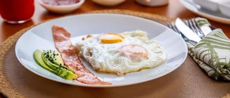 A breakfast plate with eggs, avocado, and bacon, a glass of red juice, and a white mug on a table.