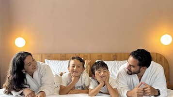A family of four in a bedroom with a wooden headboard, white bedding, and two wall-mounted lights.