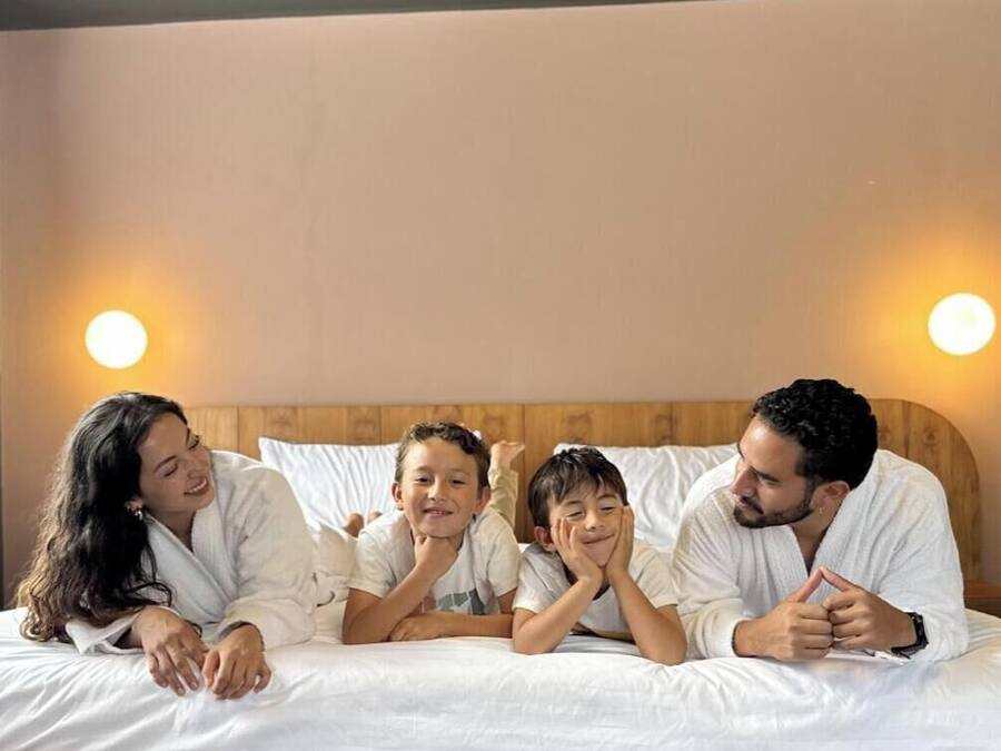A family of four in a bedroom with a wooden headboard, white bedding, and two wall-mounted lights.