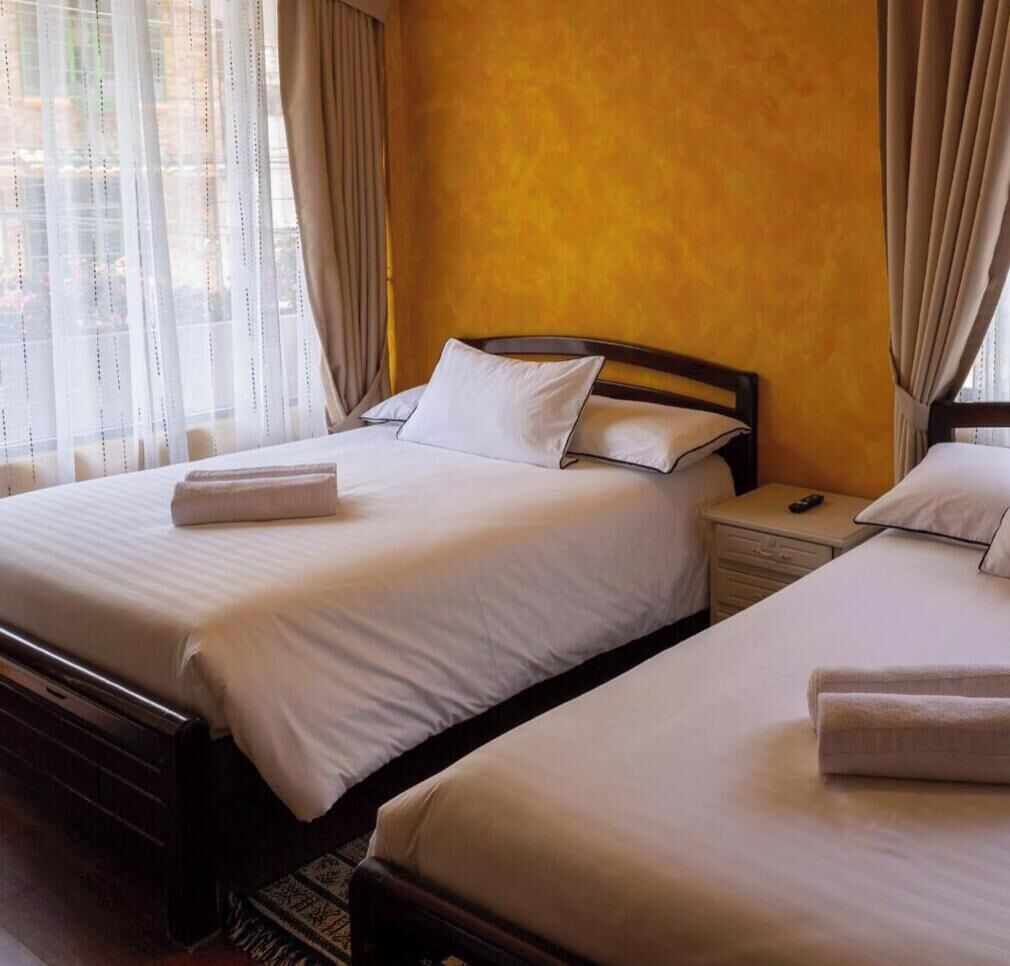 A hotel room with two beds, a wooden headboard, a bedside table with a remote control, and sheer curtains.