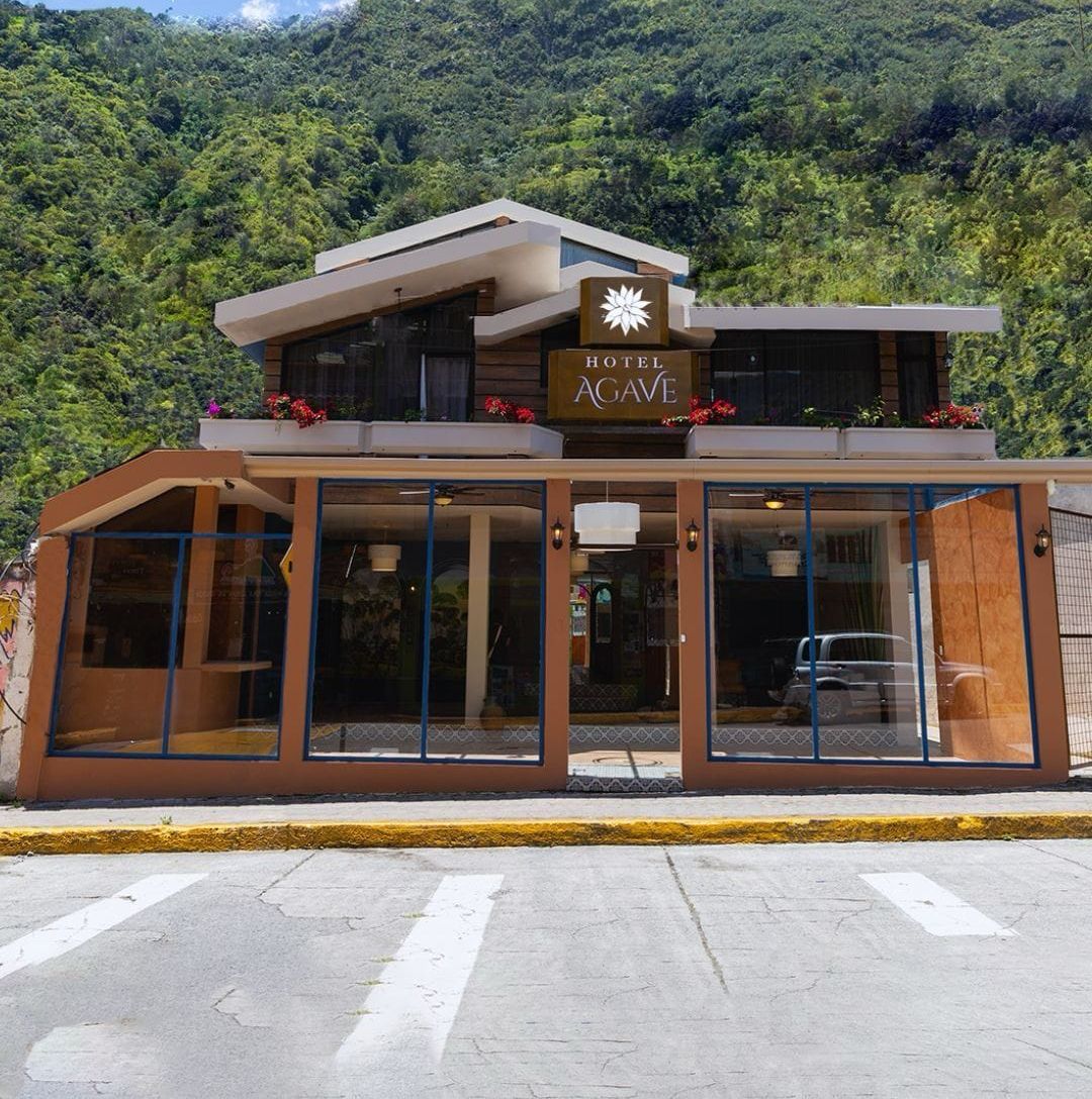 Un hotel moderno con un letrero que dice "HOTEL AGAVE" y el eslogan "toda una experiencia". El edificio tiene grandes ventanales y un techo plano.
