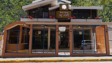 Un hotel moderno con un letrero que dice "HOTEL AGAVE" y el eslogan "toda una experiencia". El edificio tiene grandes ventanales y un techo plano.