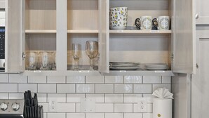 Una cocina con salpicadero de azulejos blancos, estufa y armarios abiertos que contienen cristalería, copas de vino, tazas de café y platos.