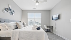Un dormitorio moderno con una cama grande, ventilador de techo, televisor de pantalla plana y ventana con vista a edificios.