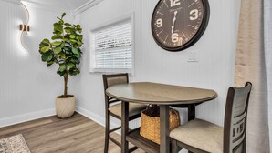 Comedor con una mesa redonda, dos sillas, una planta en maceta, un reloj de pared y una ventana con persianas.