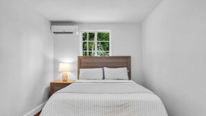 Un dormitorio con cama, mesita de noche con lámpara, unidad de aire acondicionado y ventana con vista a follaje.