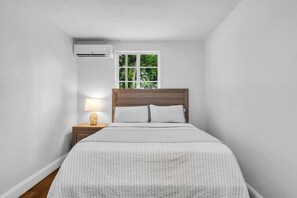 A bedroom with a bed, a nightstand with a lamp, an air conditioning unit, and a window with a view of greenery. - Cozy Private Cottage, Minutes to Anna Maria Island (Bradenton)