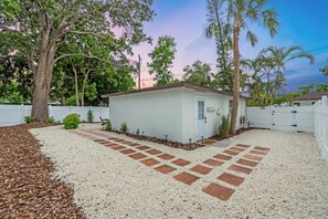Une petite maison à un seul étage, à l’extérieur blanc, entourée d’une clôture blanche, et une allée en gravier bordée de tuiles rouges.