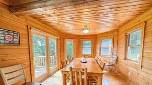 A wooden cabin with a long dining table, multiple chairs, and large windows offering views of the surrounding forest.