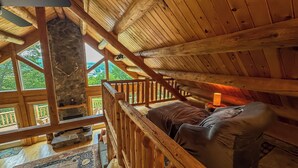 A cozy cabin with a loft bedroom featuring a wooden ceiling, a stone fireplace, and a view of the outdoors.
