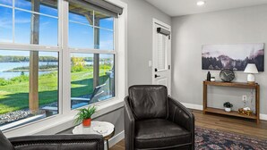 A living room with a large window overlooking a lake, a leather chair, a small table with a plant, and a console table with a clock and lamp. - RiverWest Kayak View -Hot Tub - Riverview-Dog Friendly (Duluth)