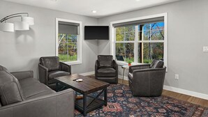 A modern living room with grey walls, a large window with a view of trees, a flat-screen TV, and leather furniture. - RiverWest Kayak View -Hot Tub - Riverview-Dog Friendly (Duluth)