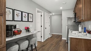 A modern kitchen with wooden cabinets, a white countertop, and a stainless steel refrigerator. There are three bar stools and framed floral artwork on the wall. - RiverWest Kayak View -Hot Tub - Riverview-Dog Friendly (Duluth)