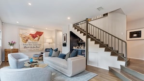 Ein modernes Wohnzimmer mit einem weißen Sofa, einem grauen Sessel und einem Couchtisch. Eine Treppe mit Metallgeländer führt zu einem dunklen Raum.