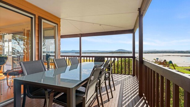 A balcony with a glass table and chairs, overlooking a beach and water body.