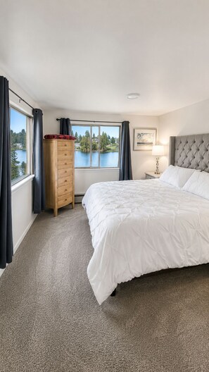 Ein Schlafzimmer mit einem großen Bett, einer Holzkommode, einem Nachttisch, einer Lampe und Seeblick durch ein Fenster.