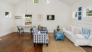 A spacious living room with a high ceiling, a dining area with a table and chairs, a blue and white checkered sofa, a white couch, and a television mounted on the wall.