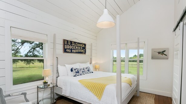 A bedroom with a bed, two bedside tables, a hanging light fixture, a window with a view of a green field, and a wall sign.