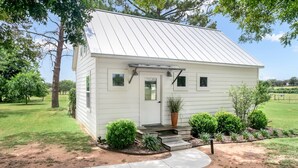 A small, single-story house with a white exterior and a metal roof. It features a front porch with steps and a potted plant.