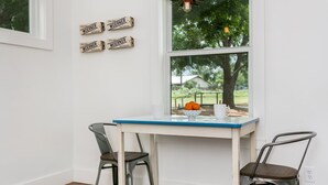A small dining area with a white table and two chairs, a bowl of fruit, and a window with a view of trees and a house.