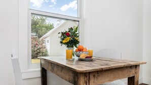 Ein Holztisch mit einer Blumenvase und einer Obstschale, Gläsern Orangensaft und einem Stuhl mit gemustertem Sitzkissen.