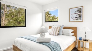 A bedroom with a large bed, two windows with views of greenery, a bedside table with a lamp, and a framed picture on the wall.