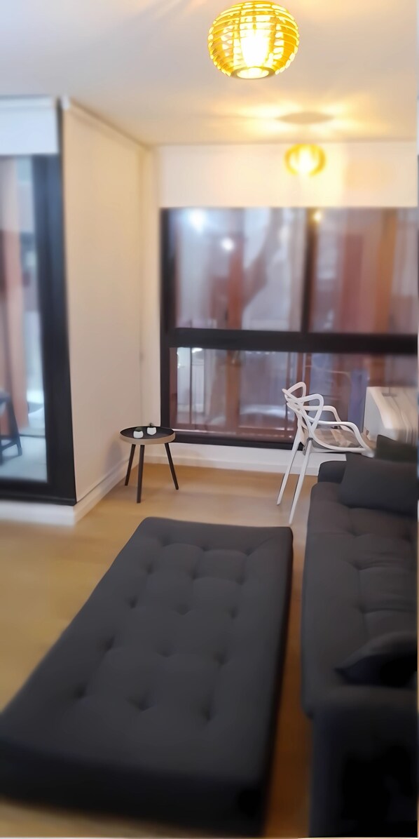 Una sala de estar moderna con un ventanal grande, un sofá negro, una mesita pequeña y una lámpara colgante.