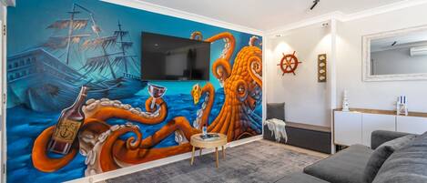 Un salón con un mural de un barco y un pulpo, un televisor de pantalla plana, una pequeña mesa redonda y un sofá gris.