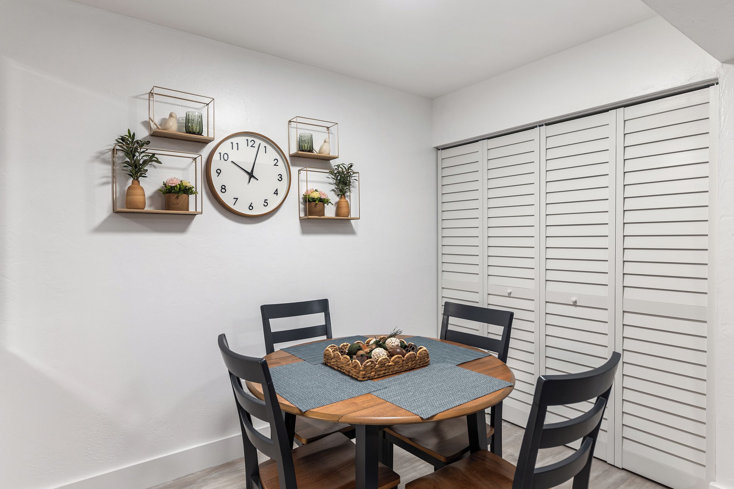 A dining area with a round table, four chairs, a clock, and three wall-mounted shelves with plants.
