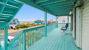 Une véranda en bois de couleur turquoise, avec une seule chaise, donnant sur une plage bordée de maisons et traversée par une route.