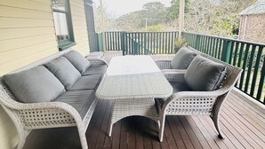 A covered outdoor seating area with a wicker sofa and table, wooden deck flooring, and a view of trees and a building in the background. - 3BDR Blue Mountains Hideaway in Central Katoomba (Katoomba)