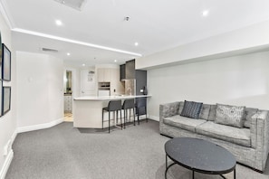 A modern apartment with a grey sofa, a small round table, and a kitchen area with a bar counter and stools. - KozyGuru | Sydney CBD | Skyline View Sleek Studio (Sydney)