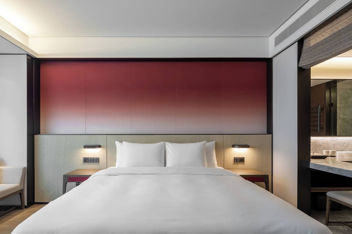A modern hotel room with a large bed, bedside tables with lamps, a red accent wall, and a view of the bathroom.