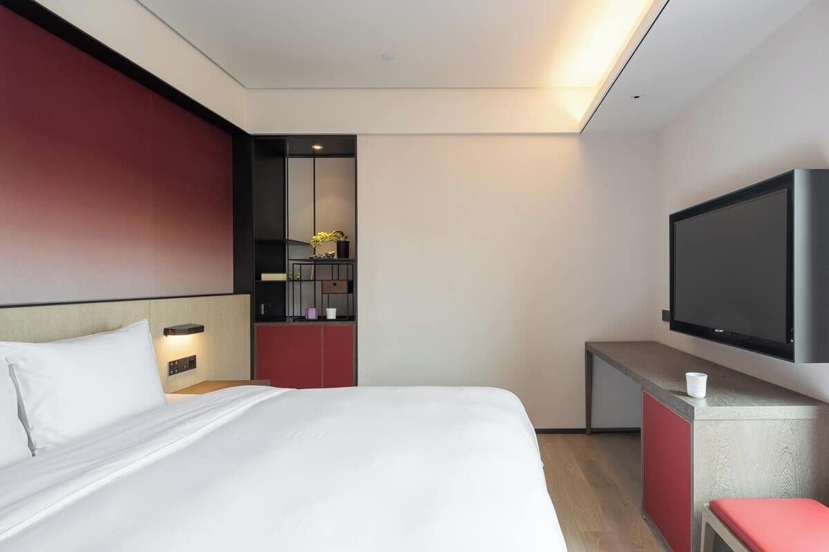 A hotel room with a large bed, a TV mounted on the wall, a small desk, and a red bench.