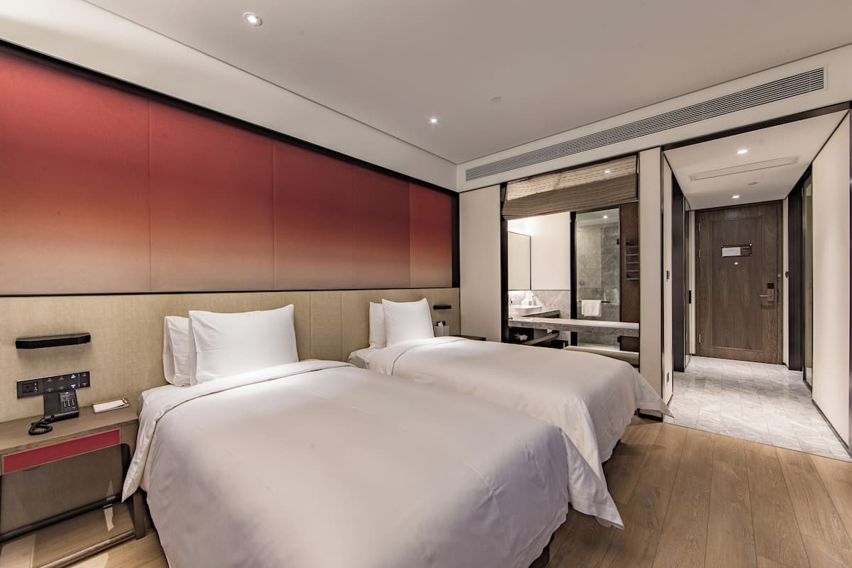 A hotel room with two beds, a bedside table with a phone, and a mirror above the sink.