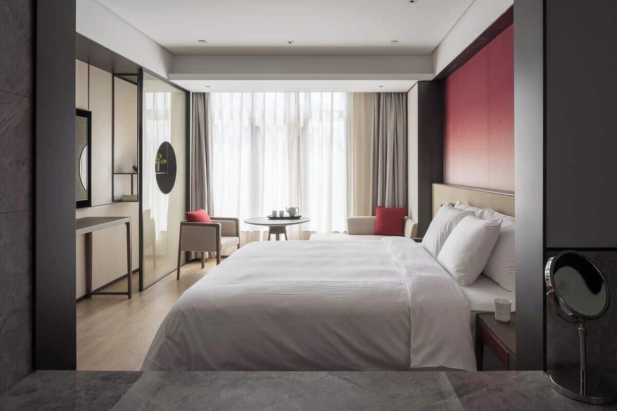 A modern hotel room with a large bed, a desk, a chair, and a small table.
