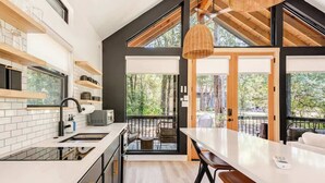 A modern kitchen with white countertops, black cabinetry, and a large island with bar stools. There are wooden shelves above the counter and a large window providing a view of the outdoors. - Relaxing Ranch, Friendly Animals, Modern Stay (Cedar Creek)
