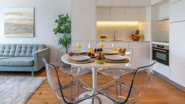 Una cocina moderna con armarios blancos, un comedor con mesa y sillas de vidrio, y un sofá gris en la sala.