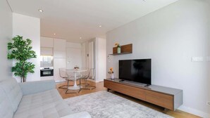 Una sala moderna con un sofá gris, un televisor sobre un mueble de madera, una mesa de comedor con sillas y una planta grande.