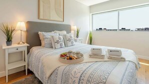 Un dormitorio ordenado con cama, mesitas de noche, una ventana con vistas a la ciudad y una obra de arte enmarcada en la pared.