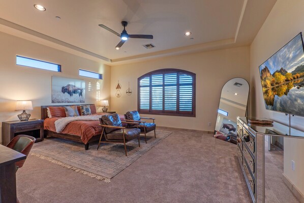 A spacious bedroom with a large bed, a desk, a chair, a ceiling fan, and a large window with blinds.